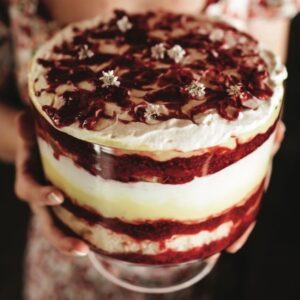 Hands holding trifle showing bright layers of raspberry and cream filling, topped with raspberry coulis and edible flowers